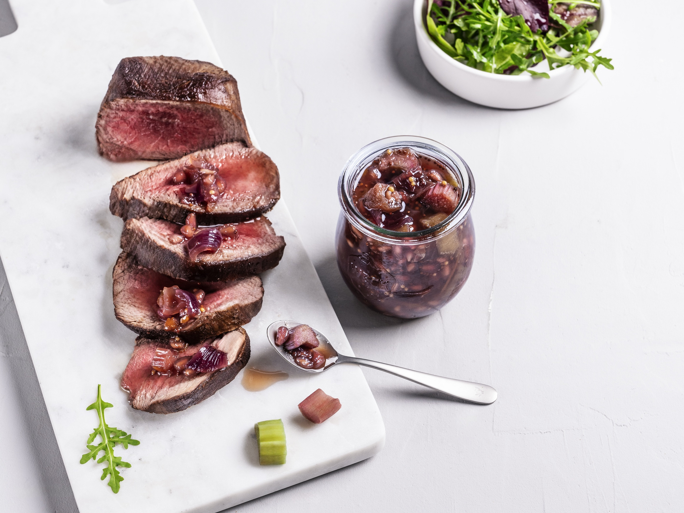 Ein Weckglas mit dunkelrotem Rhabarberchutney, daneben kurzgebratenes Rindfleisch, in Scheiben geschnitten und mit Chutney betträufelt. Im Hintergrund eine Schüssel mit grünem Wildsalat.